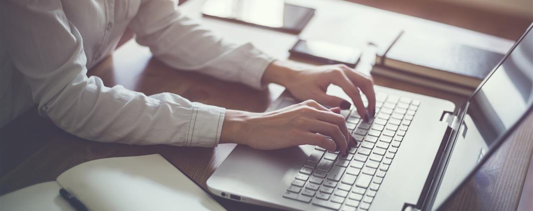 Person writing text on laptop with notebook open next to them on desk